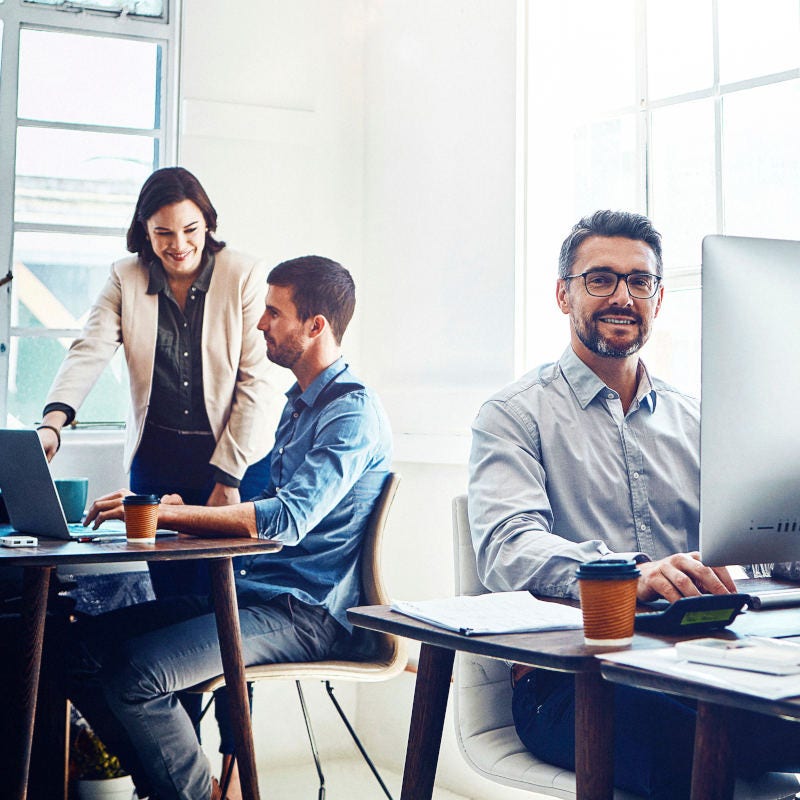 Hombre sonriendo en su escritorio trabajando en computador. Tras él se encuentran dos compañeros conversando